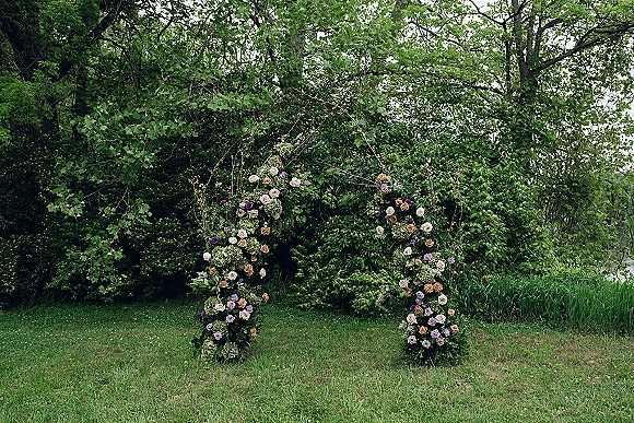 Wedding ceremony arch with an asymmetrical rose and greenery floral frame on a grassy lawn, with trees and water behind