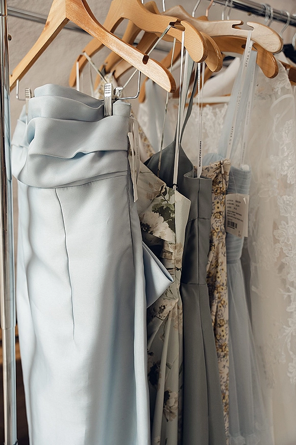 Bridesmaid dresses in dusty blue bridesmaid dresses hang on wooden hangers with a lace wedding dress on a rack against a neutral wall