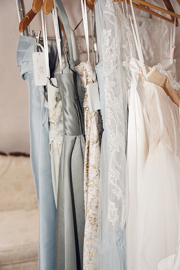 Bridesmaid dresses, dusty blue bridesmaid dresses on hangers along a clothing rack with lace veil and ribbons against a neutral wall