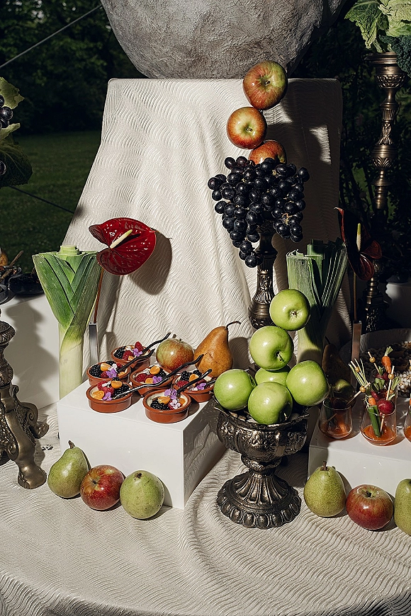 Wedding dessert table with fruit wedding dessert table styling, silver urns, pedestal bowls, and mini dessert cups against dark garden foliage.