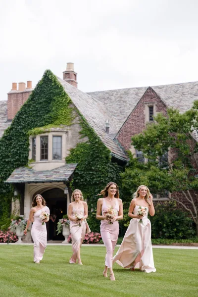 Bridesmaid portraits of bridesmaids walking photo in blush dresses, holding bouquets and heels, outside an ivy-covered brick manor lawn
