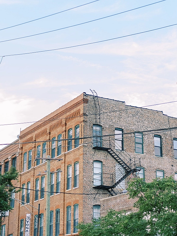 Brick building exterior with a black metal fire escape and rows of windows, framed by power lines with trees and sky behind