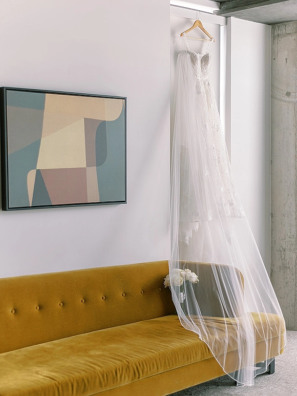 Wedding dress on hanger with a long veil, lit by window light against a white wall, with bouquet resting on a velvet sofa