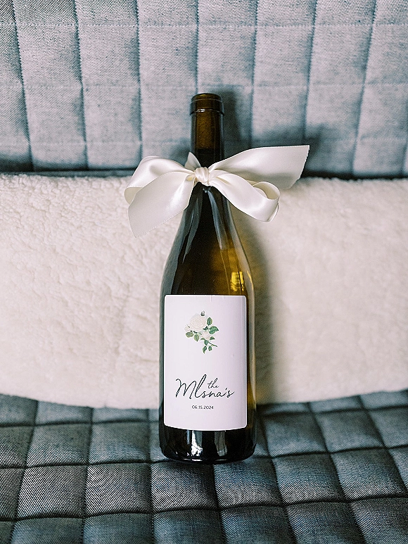 Wedding wine bottle with personalized wine label and a white satin ribbon bow, resting on a folded white towel by an upholstered chair