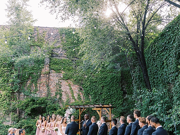 Ceremony moment at an outdoor wedding ceremony with bride and groom under a wooden arbor, officiant speaking, bridal party before an ivy brick wall