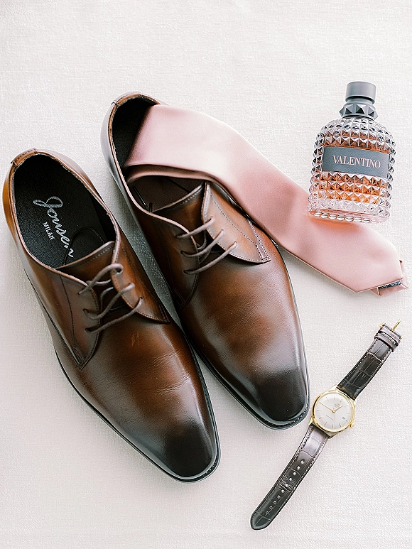 Groom accessories flatlay with brown leather dress shoes, blush pink necktie, cologne bottle, and wristwatch on light fabric surface