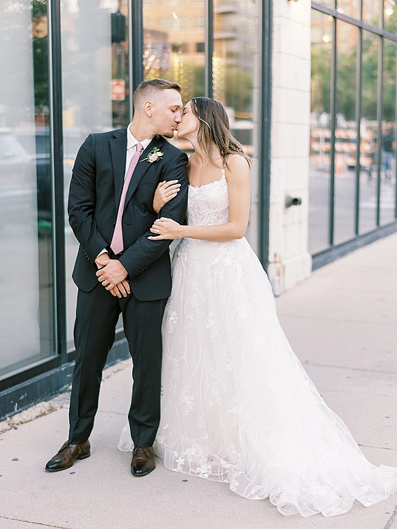 Wedding kiss portrait of bride and groom kissing by glass windows on a city sidewalk, lace gown train and blush boutonniere visible