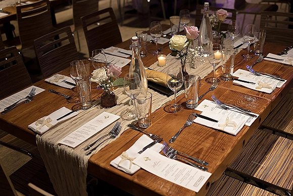Reception tablescape with rustic wedding table styling, linen runner, roses and baby's breath in glass bottles, votive candles, menus and place settings indoors