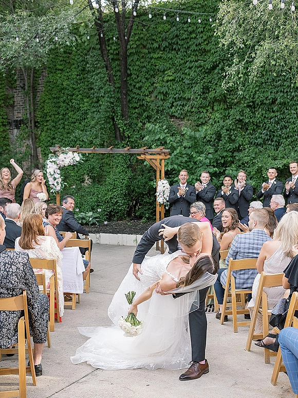 Ceremony kiss moment as groom dips bride in veil and dress, bouquet in hand, beneath floral wooden arch with string lights in ivy courtyard