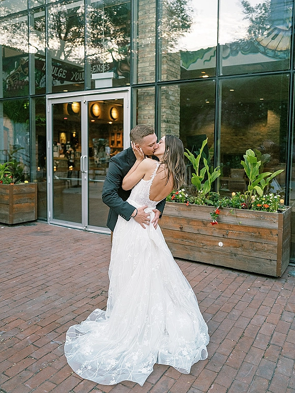 Wedding kiss portrait of bride and groom kiss on a brick walkway by a glass restaurant facade, her low-back lace gown trailing behind