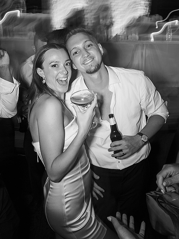 Wedding reception guests dancing on the dance floor, one holding a cocktail glass amid reception lighting and a draped wall behind them