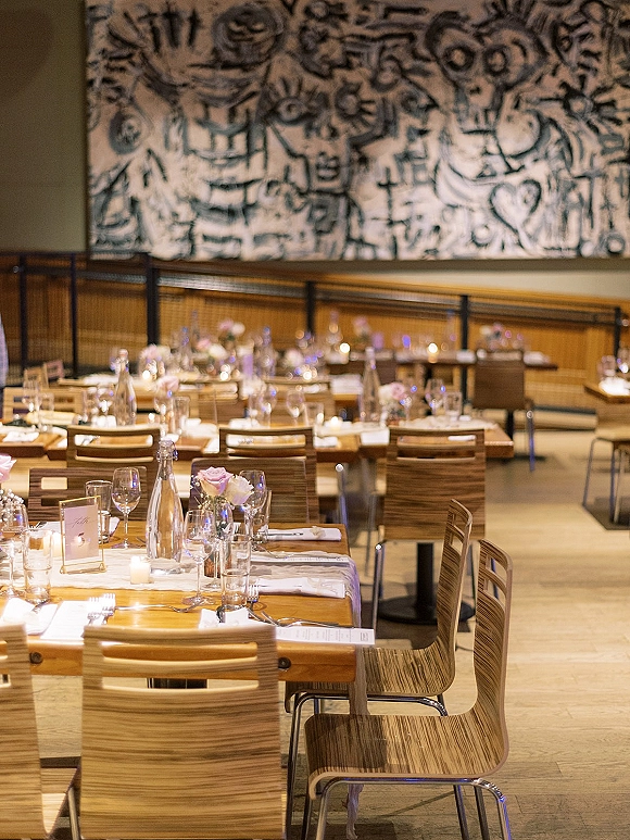 Reception tablescape with a white runner, menus, glassware, votive candles, and pink rose bud vases on a long wood table in a mural-lined hall