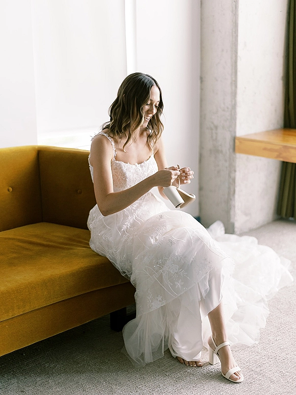 Bride getting ready unboxing bridal shoes photo while seated on a mustard velvet sofa in lace spaghetti strap gown by a large window