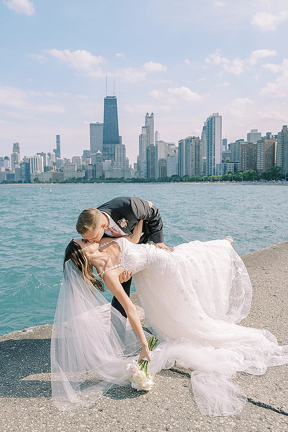 Wedding kiss portrait of groom dipping bride in lace dress and veil on a concrete pier by a lake with city skyline behind