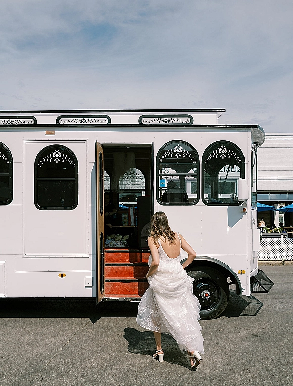 Bride portrait of a bride walking away toward a white trolley bus, showing an open-back lace tulle dress and bridal heels at the door