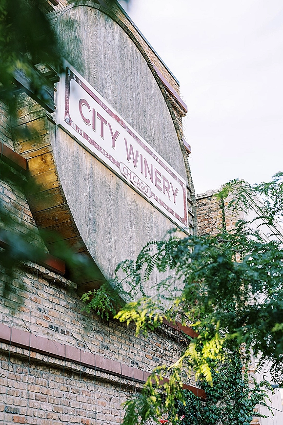 Wedding venue exterior with a wedding venue sign in painted lettering on brick facade, wood trim, and ivy vines under trees
