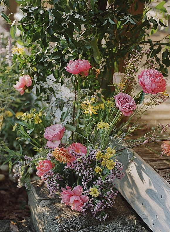 Wedding floral arrangement with peony wedding flowers in a wooden crate, featuring pink roses and yellow wildflowers on a sunlit stone ledge