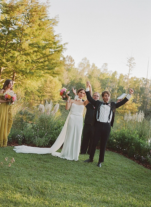 Ceremony recessional as bride and groom recessional raise their hands, bride in strapless gown and long veil on a garden lawn aisle