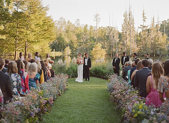 Outdoor wedding ceremony with bride holding bouquet beside groom in black tuxedo, guests seated on a lawn by a lake and trees