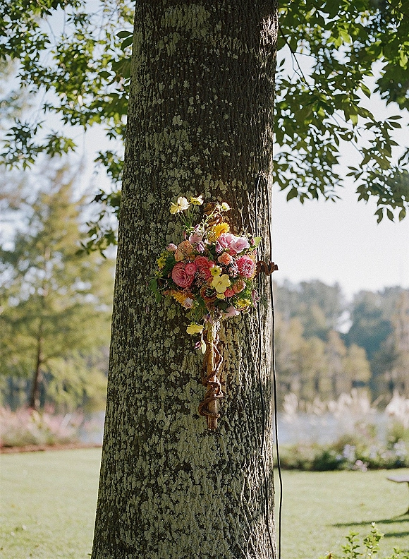 Hanging wedding bouquet tied with ribbon to a tree trunk, colorful blooms suspended on rope with a lakeside lawn and trees behind
