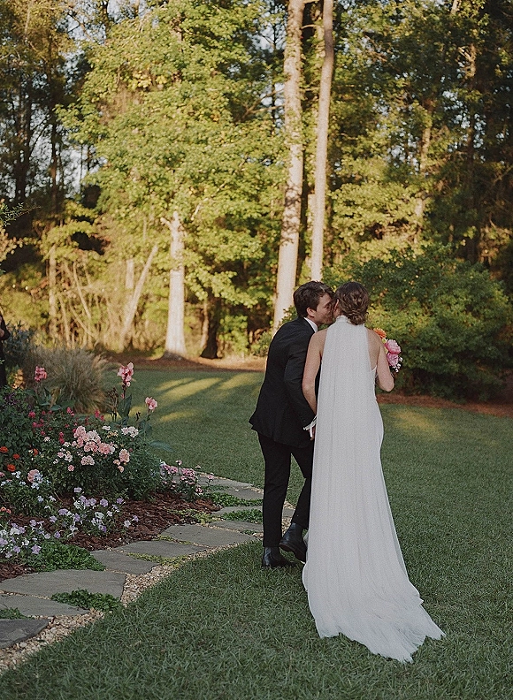 Wedding kiss as bride and groom walk hand in hand on a garden stone path, her long veil and pink bouquet trailing behind