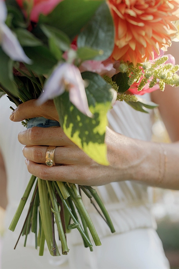 Bridal bouquet with an orange dahlia bouquet of pink blooms and greenery, blue ribbon wrap, and a wedding ring against white dress fabric