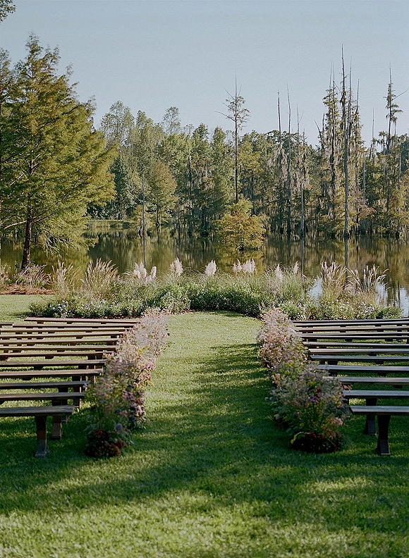 Ceremony aisle setup with wooden benches and flower-lined ground arrangements, framed by ornamental grasses on a lakefront lawn
