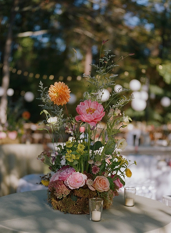 Wedding centerpiece with wildflower blooms, pink peonies, garden roses and orange dahlia on a moss base with votive candles under string lights