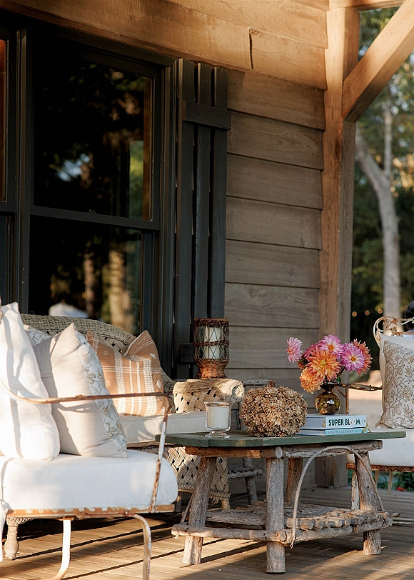 Lounge seating area with wedding lounge furniture on a wood porch, styled with pillows, rustic coffee table, candles, lantern, and bright floral centerpiece