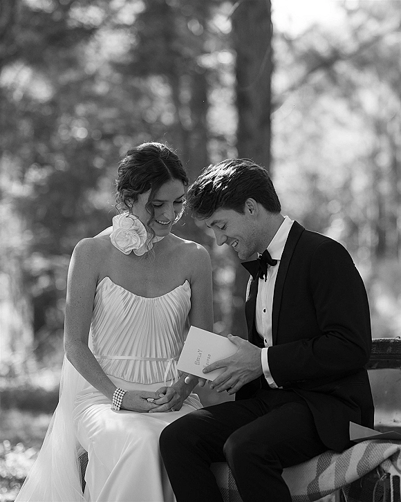 Couple portrait of bride and groom sitting on a bench as groom reads a wedding vow book, plaid blanket and sunlit park bokeh behind