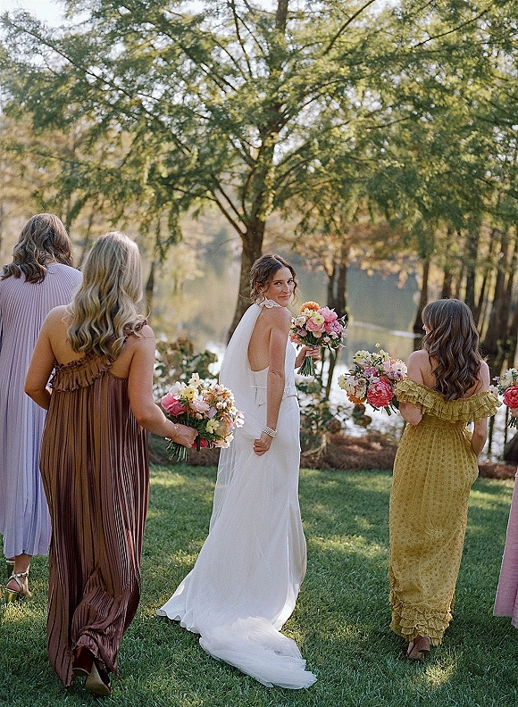 Bridal party portrait of bride with bridesmaids walking on a grassy lawn, holding bouquets, with trees and a lake behind them
