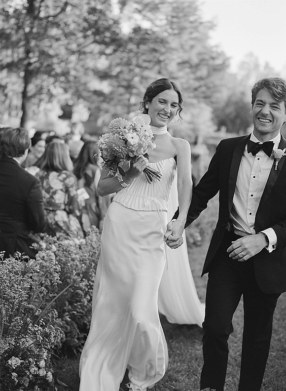 Wedding recessional as bride and groom walking hand in hand, bride holding a bouquet, smiling past guests in an outdoor garden path