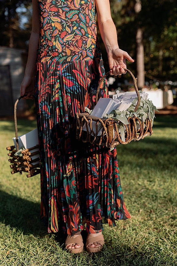 Wedding programs stacked in a woven basket with eucalyptus accents beside a guest in a floral maxi dress on a grassy lawn outdoors