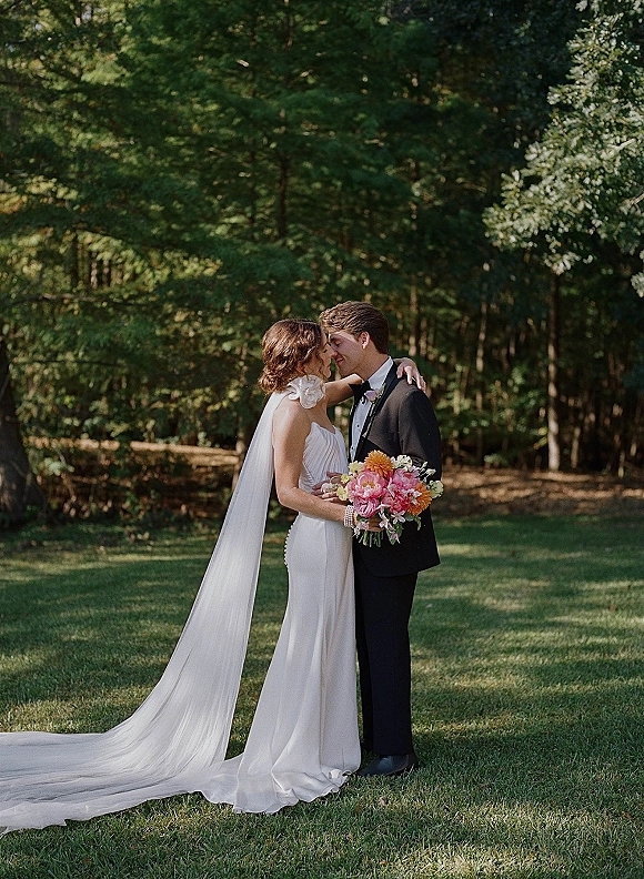 Wedding couple portrait of bride and groom kiss in natural sunlight on a lawn by forest trees, bride holding bouquet with long veil
