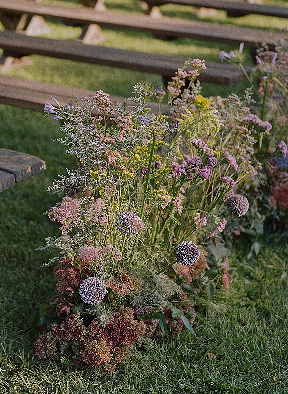 Aisle floral arrangements of meadow blooms and greenery with purple allium accents lining a grass lawn ceremony with wooden benches
