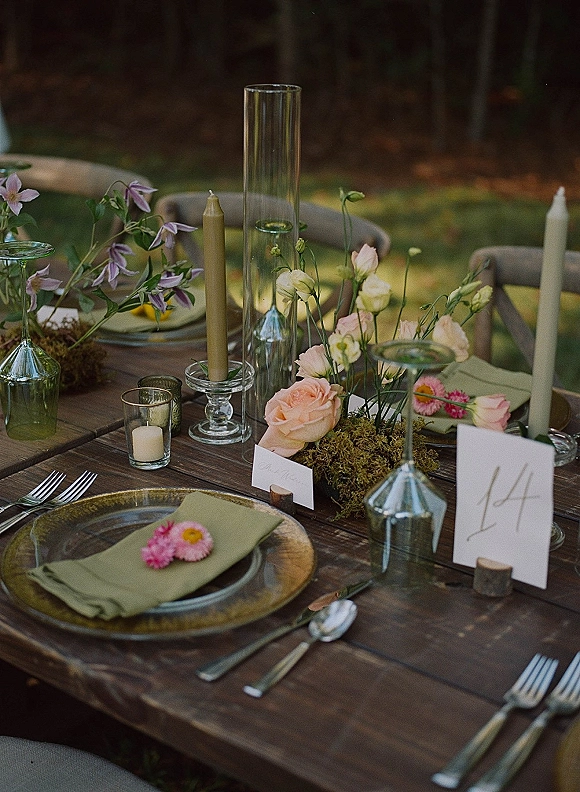 Reception tablescape with rustic wedding table setting on a wood dining table, moss and floral centerpiece, taper candles in glass holders on a lawn under trees