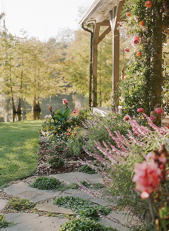 Wedding floral installation with outdoor ceremony florals, climbing vines and roses cascading from porch pillars by a sunny lakeside lawn