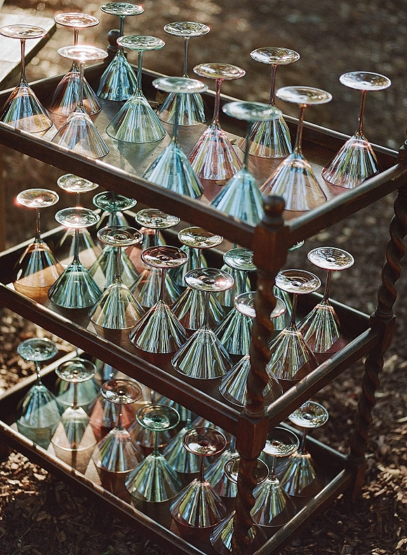 Champagne coupe display with colored coupe glassware arranged on a wooden bar cart in dappled sunlight outdoors