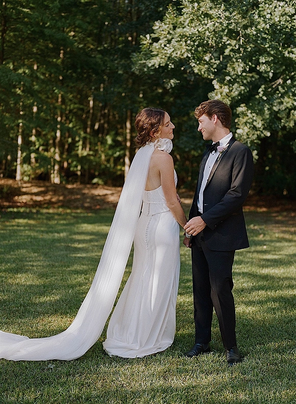 Couple portrait of bride and groom holding hands, her long veil trailing on grass beside his black tuxedo at a sunlit forest edge