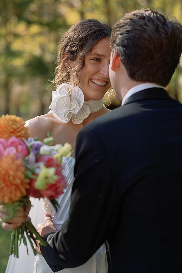 Wedding couple portrait of bride smiling at groom, holding a colorful bouquet in an outdoor garden with lush greenery behind them