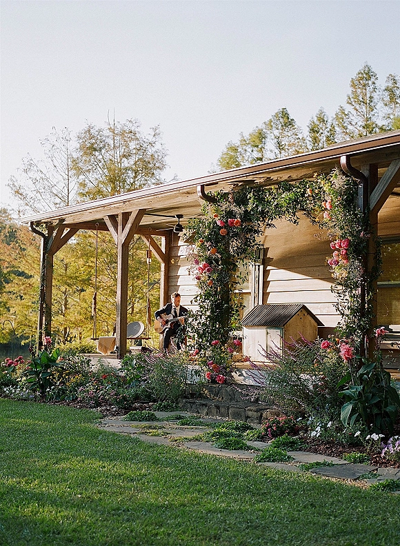 Wedding ceremony music by a wedding guitarist in a suit playing acoustic guitar on a cottage porch with a floral arch and greenery garland