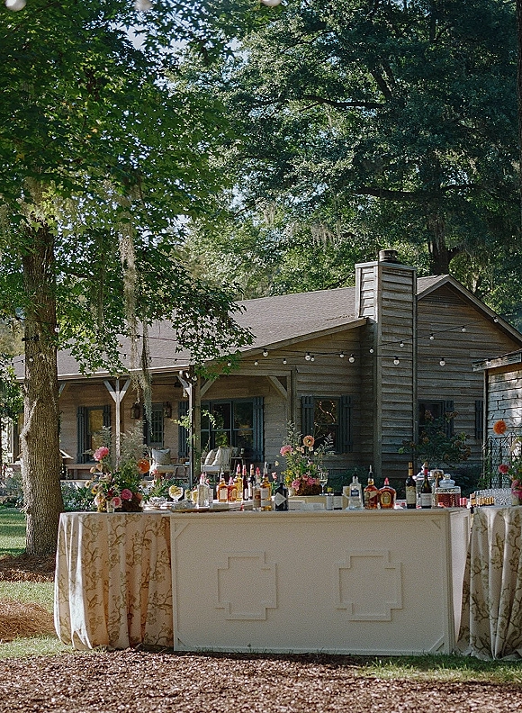Outdoor wedding bar with liquor bottles and glassware on a patterned tablecloth, floral accents and string lights by a cabin porch