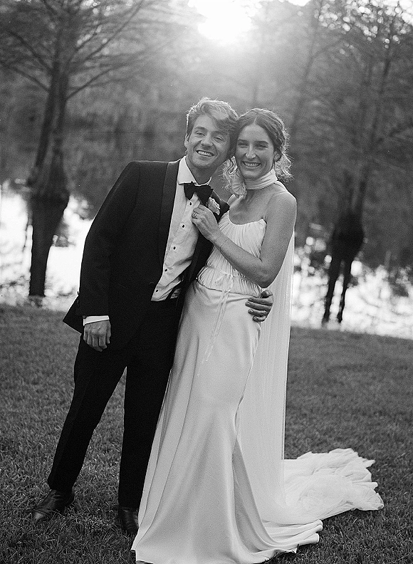 Couple portrait of bride and groom smiling as they embrace by a tree-lined lake, her veil flowing over a strapless dress in sunlight