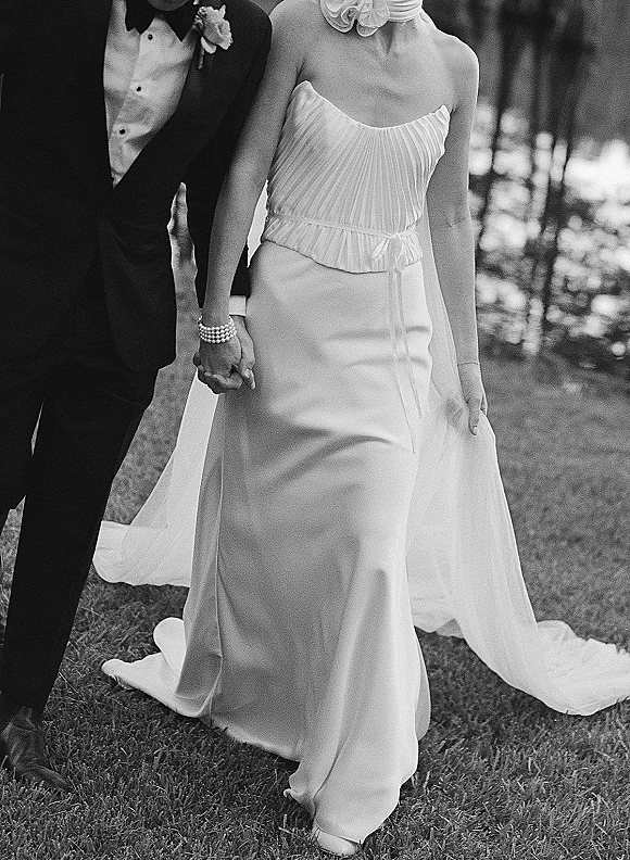 Couple portrait of bride and groom holding hands in a black and white wedding photo, her strapless dress and long veil on a lawn with trees