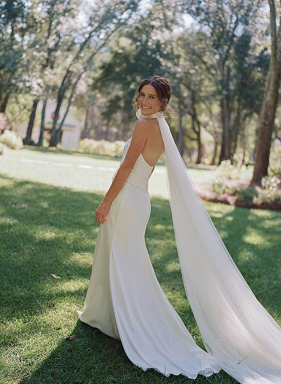 Bridal portrait of a bride looking back in a halter neck, low-back wedding dress with a long veil on a sunlit garden lawn