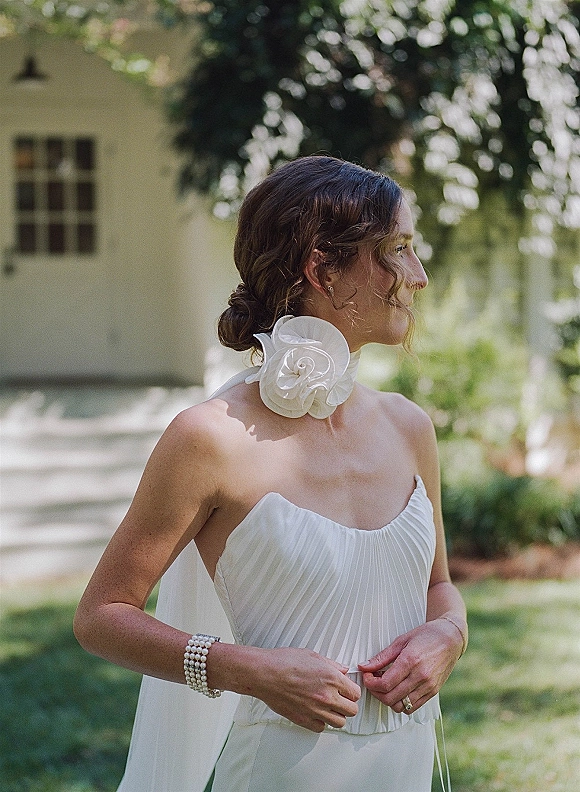 Bridal portrait in a strapless wedding dress, wearing a floral neck scarf and veil, standing on a sunlit garden lawn near trees and a white building