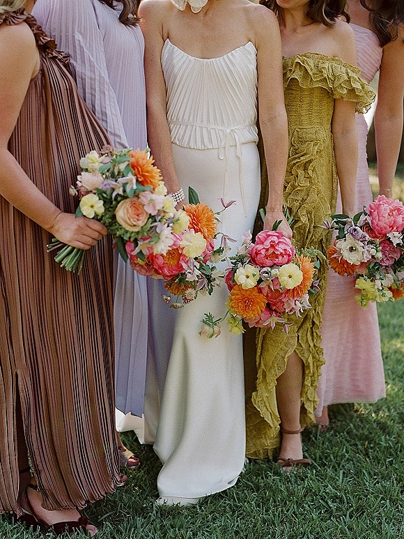 Bridesmaid group photo with bridesmaids holding bouquets in mismatched pastel dresses and colorful flowers, standing together on a grass lawn
