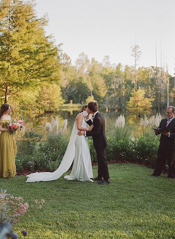 Ceremony kiss as bride and groom embrace, her long train and veil flowing beside yellow bridesmaid holding bouquet at lakeside lawn altar