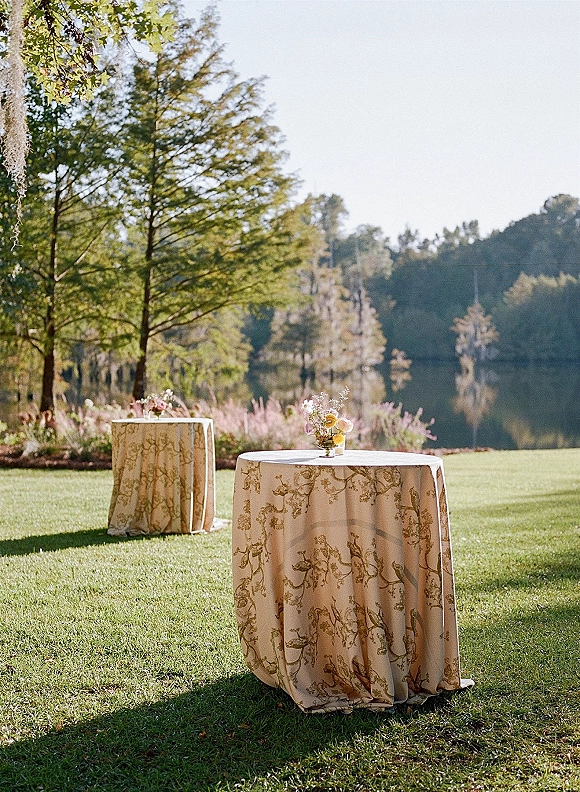 Cocktail table decor with patterned linens and small floral bud vases on round tables set on a lawn by a lake and trees