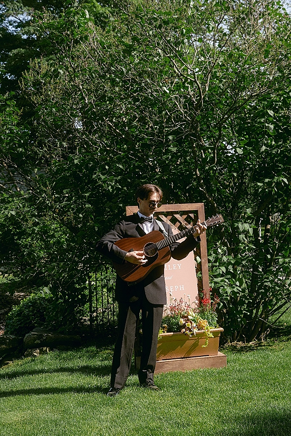 Wedding musician playing acoustic guitar in a tuxedo and sunglasses beside a floral welcome sign on a green garden lawn